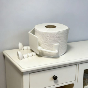 White toilet paper roll on a decorative novelty holder with a small door and curved lever, on a white dresser.